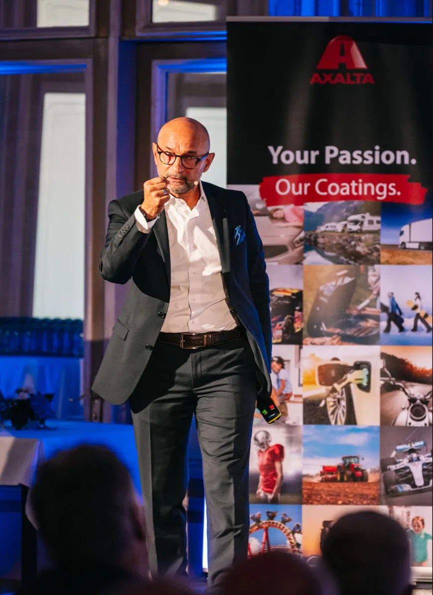 Enzo Santarsiero spricht mit Headset-Mikrofon auf einer Bühne, daneben ein Axalta-Rollup „Your Passion. Our Coatings.“