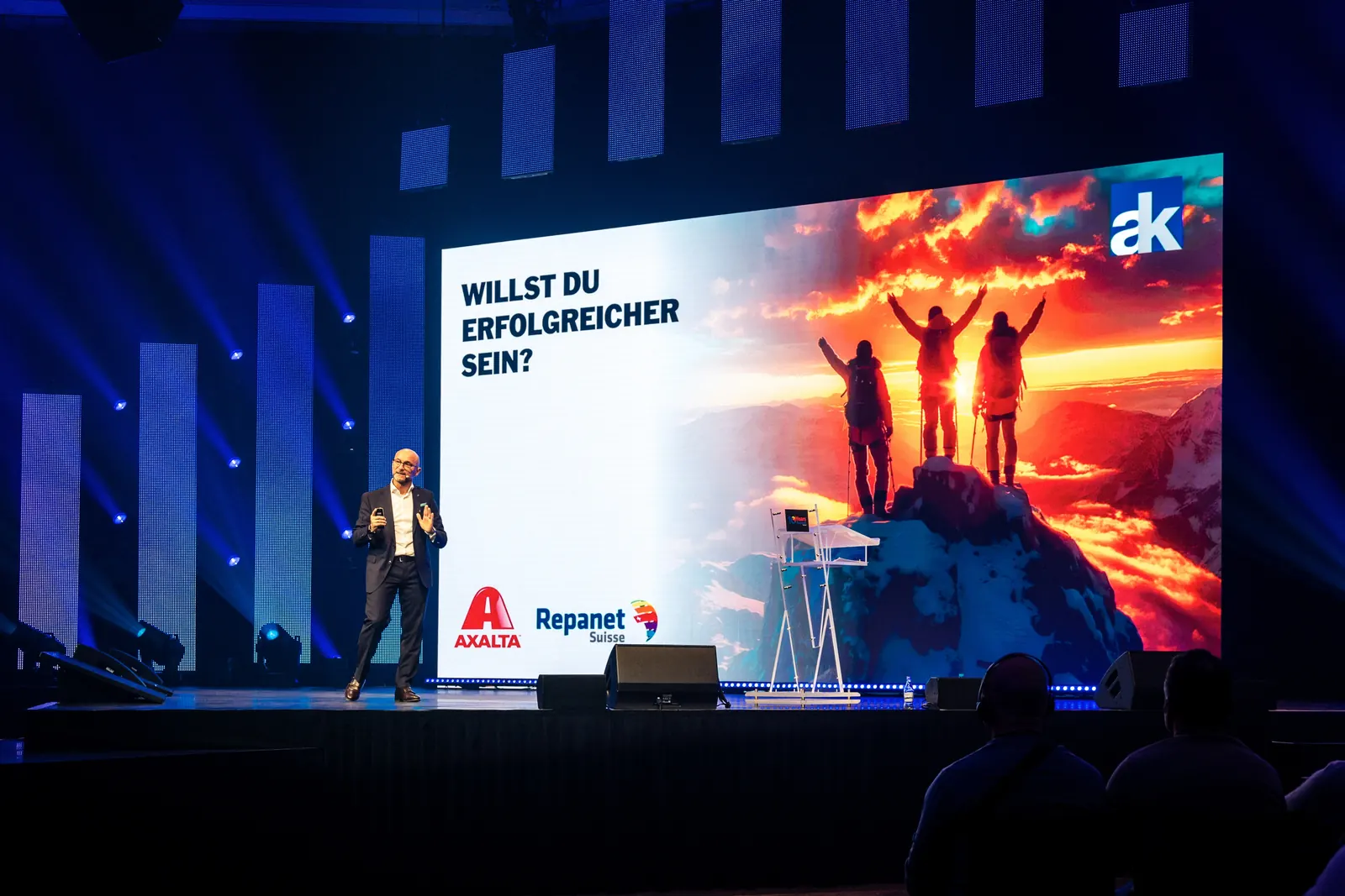 Enzo Santarsiero auf einer grossen, blau beleuchteten Bühne vor einer Leinwand mit der Frage „Willst du erfolgreicher sein?“ und den Logos von Axalta und Repanet Suisse